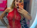 NicolSNaya with blonde hair, red lace-up bodysuit, showing her tits, holding an orange phone, purple lipstick, tattoos on arms, pink nails, in a mirror selfie.