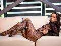 IsadoraReyes is lying on a white couch, wearing a black lace bodysuit that exposes her tits and pussy through the pattern. Her legs are stretched out, looking sexy.