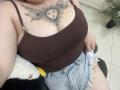 DiLaria's large boobs in a brown tank top, showing a sun face tattoo between them. She's wearing frayed denim shorts, sitting with a white and black blanket.