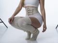 Destinis is squatting, wearing a white crop top, white shorts, beige belt with buckle, and chunky cream knit socks. Her blonde hair is visible, showing her back.