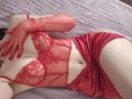 Rozalin in red lace lingerie and matching satin shorts, with red mesh gloves, lying on a textured gray bed. Her tits are small, her ass is not visible.