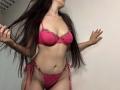 Tisia with long dark hair, pink lace bra with floral embroidery, pink lace panties with side ties, standing, hands in hair, smiling, showing her tits and pussy.