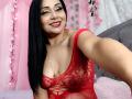 Zaria69 with long black hair, red lace top showing her tits, smiling at the camera, extending arm for selfie, pink and white background, pink fairy lights, and tattoo.