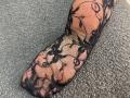 Tattiya's lace-covered cock, featuring black floral patterns, is laid out on a textured gray surface. The lace is sheer, revealing a tan, circumcised, erect penis underneath.