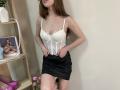 StellaNoir has pale skin, long brown hair, and small tits in a white lace corset, black mini skirt, and no pants, standing with hands on skirt, in a room.