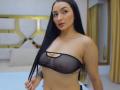 PamelaLiss with long black hair, wearing a sheer black bra, showing small, perky tits with visible nipples, standing in a room, holding her hair, tattoo on right side.
