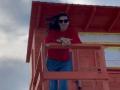 Nalini, con lunghi capelli neri, un maglione rosso, pantaloni blu e occhiali da sole neri, appoggiata a una struttura di legno rossa, sorride, con il sedere leggermente visibile, in piedi contro un cielo nuvoloso..