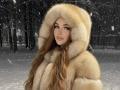 BryannaCrecelius with long brown hair, wearing a fluffy tan fur hood, snow falling, winter forest, soft pink lips, slight smile, sexy, attractive, white snow on fur.