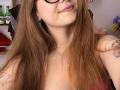 KarKutie with long brown hair, black glasses, and subtle smile, showing a tattoo on her right arm, and a necklace, with visible bare chest and pink top.