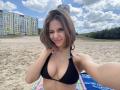 MilagrosEdmisten with shoulder-length brown hair, smiling, wearing a black bikini that reveals her cleavage, standing on a sandy beach with colorful buildings and a cloudy sky in the background. Beach towel under her.