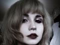 EmelyDilsaver has pale skin, dark eyeliner, shiny pink eyeshadow, and dark purple lipstick. Her blonde, messy hair frames her freckled face in a slightly blurred, dark photograph.