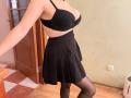 EmmaNoa con grandi seni, indossa un reggiseno nero, una gonna nera e calze nere, è in piedi in una stanza, con una porta di legno sullo sfondo, e ha un tatuaggio sul lato destro della gabbia toracica..