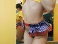 O bumbum nu de VioletaNova é visível em um reflexo no espelho, usando uma minissaia roxa e vermelha brilhante com babados, com uma blusa amarela e uma tanga vermelha à mostra, em um quarto amarelo..