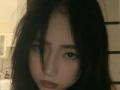 MargeretEskuchen has pale skin, dark messy hair, black eyebrows, dark eyeliner, and a lip piercing, looking down with a slightly pouty expression in a dimly lit room.