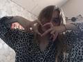 BessBetak with wavy blonde hair making a heart shape with her hands, wearing a leopard print shirt, has pink nails, and a black wristband, in a dimly lit room.