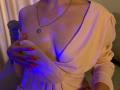 Anshante's tits are exposed in a pink dress with a deep V-neck, her hand covering one with red nails, wearing a silver necklace with a square pendant.