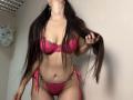 Tisia with long brown hair, wearing a pink lace bra and matching panties, holding her hair, showing her tits and pussy. Her skin is light, she's standing in a room.