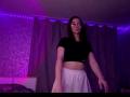 SunnyKlusman with long brown hair, wearing glasses, black crop top, and white skirt, standing in purple-lit room, arms slightly raised, looking at camera.