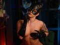 LexiSolene with black cat mask, fingerless black latex gloves, and tattoos, touching her lips, topless, showing her tits, in a dimly lit room.