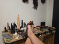 BeccaVanBoa's legs with brown high heels on a cluttered desk, featuring a large dildo, black sex toys, scissors, and various BDSM items, with black feathers hanging above.