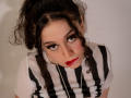 KendraSimons with curly dark hair in a ponytail, wearing a striped black and white shirt, looking up with red lipstick, white string around her neck, and heavy eyeliner.