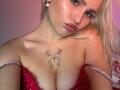 BaeonLive with blonde hair, red lipstick, and a butterfly tattoo between her tits, wearing a sparkly red dress, looking at the camera with a slight pout.