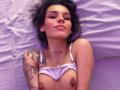 LenaBranch lies on a bed with purple sheets, topless, wearing a white bra pulled down, showing her tits. She has tattoos, freckles, and a "FEM" tattoo on her neck.