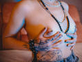 JadeMilleer's topless with hands covering her tits, adorned with nipple piercings and black nail polish. She's wearing beaded necklaces and a spiked bracelet, showing tattoos on her belly.
