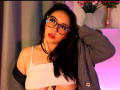 SusanMonroy with glasses, black hair, and red lipstick, wearing a white crop top, gray jacket off, exposing her small tits, hand in hair, pink and blue light reflection.