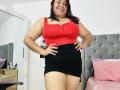 KarlaWild with curly red hair, wearing a red tank top showing her big boobs and a black skirt, standing with hands on hips, smiling confidently. Bedroom setting.