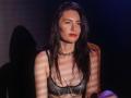IngaDrein with wet black hair, red lipstick, chain necklace, and sparkly black bra, sitting, shadowed, striped light on her tits, and a chain across her chest.