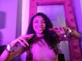 SophiaLuthor with curly dark hair, smiling topless, wearing pink-tufted mirror frame, purple lighting, and chunky silver bracelets, holding her tits with pink-painted nails.