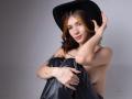 GretaHarris, topless, wearing black hat and black leather pants, holding knees, tattoos on arms, wavy brown hair with orange highlights, slight smile, gray background, hands on hat.