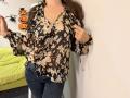 Tilliana vestindo uma blusa floral marrom, jeans azuis e unhas cor-de-rosa, segurando as bordas da blusa, com cabelos castanhos longos, mostrando os seios, em pé em frente a uma parede branca..