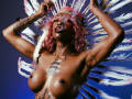 Topless DaniaMarcell with pink curly hair, feather headpiece, dark skin, tattoos, large boobs, arms raised, blue background, white feathers, pink headband, shiny skin, confident expression, sexy.