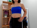 Steisii with a blue tube top showing her tits, black skirt, and hand on her hip, reflected in a mirror, wearing a necklace, in a room with wooden shelves.