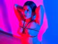MelodyRoss with arms behind head, wearing a black strappy harness, exposing her tits, lit by pink and blue neon lights, standing on wooden floor.