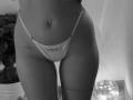SugarrX's lower belly and hips, wearing a thin, white, string thong, showing a belly button piercing and tattoos on her right side and left thigh. Black and white photo.