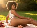 RoldanStefy, a dark-skinned woman with a big afro, sits on a pink blanket, wearing white lace lingerie, showing her ass, with grapes beside her.