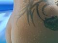 FrenchieLilieFleur's wet, tattooed ass with droplets of water, showing a green frog tattoo and dark, leafy lines. The photo focuses on the right buttock.