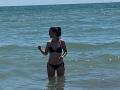 LanyJolene, with dark hair in a bun, wearing a black bikini, stands in shallow blue-green water, looking to the side with her right hand raised.