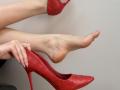 Tattiya's bare feet with a pinkish tone, one foot in a red, quilted, high-heel shoe, the other foot raised, holding the shoe with her hand.