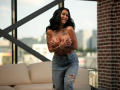 NyraNoble, topless, with tattoos, covers her big tits with hands, wearing ripped blue jeans, smiling, black wavy hair, standing in a bright room with a brick wall.