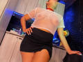 LaurenHills in a sheer white top showing her tits, black skirt with a thigh slit, red choker, hands on hips, leaning back in a kitchen with blue lights.