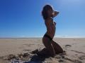 Aiyana kneels on a sandy beach, topless with small tits, wearing black panties, sunlight on her skin, blue sky, her hand in her wavy brown hair.