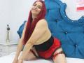 CamilaLux with red hair, smiling, topless, wearing black lace garter, red skirt, and black corset, kneeling on bed, blue tufted headboard, pale skin, looking at camera.