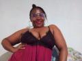 CavyQueenForU with dark skin, glasses, and hoop earrings, wearing a black lace and pink babydoll, showing her big boobs and smiling. She's sitting with one hand on her hip.