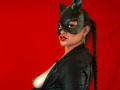 AdalinaVera with cat mask, black leather outfit, and braided ponytail, showing her tits, red lipstick, and gold hoop earrings, against a solid red background.