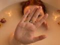 KatiRokisky's red-haired face is partially submerged in a bathtub, with her right hand, showing a ring, raised in front. Water and roses are in the background.
