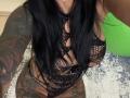 AndraD with black fishnet lingerie, showing large tits, tattoos on arms and thighs, long black hair, and a green inflatable in the background. AndraD's lower body is partially visible.